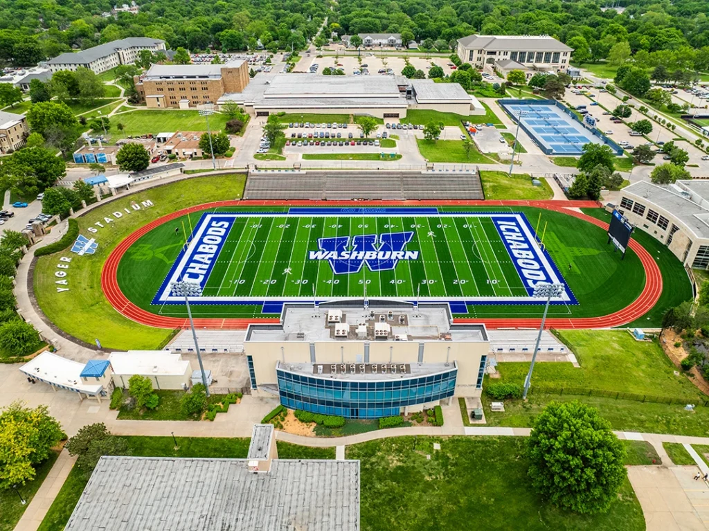Aerial view of Washburn Yager Stadium turf and its Topeka campus, showing the green GAME ON field with navy ICHABODS end zones, the Washburn W at midfield and the surrounding red track