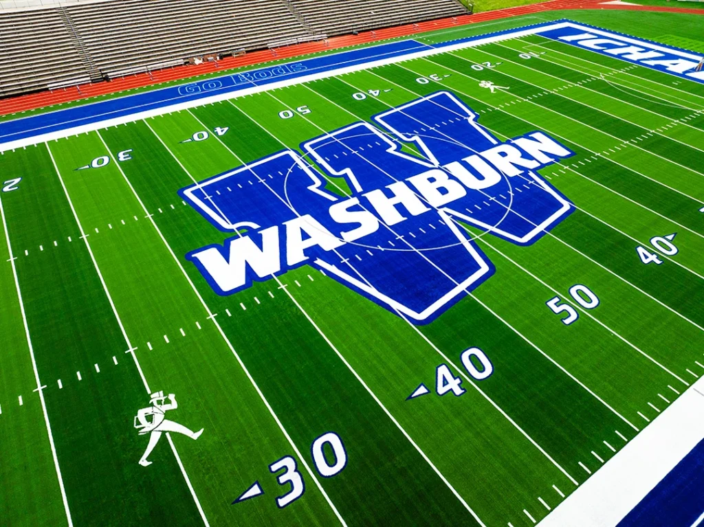 Close aerial of the huge Washburn W logo at the 50-yard line, tufted into the green GAME ON turf beside the white Ichabod mascot