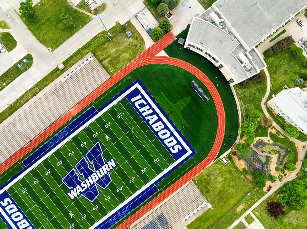 Overhead drone view of the Washburn Ichabods football field, with the navy WASHBURN wordmark and W at midfield, Go Bods player boxes, white Ichabod mascot and the ICHABODS end zone