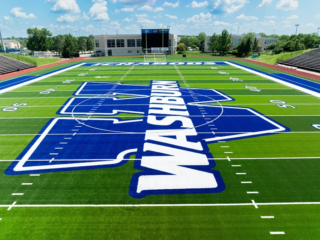 Aerial view down the length of Yager Stadium field from behind the navy WASHBURN end zone, showing the midfield W, grandstands and campus beyond