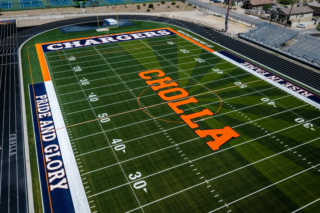 Close aerial view of the Cholla High School turf football field, showing the orange “CHOLLA” midfield logo and a navy “PRIDE AND GLORY” sideline banner