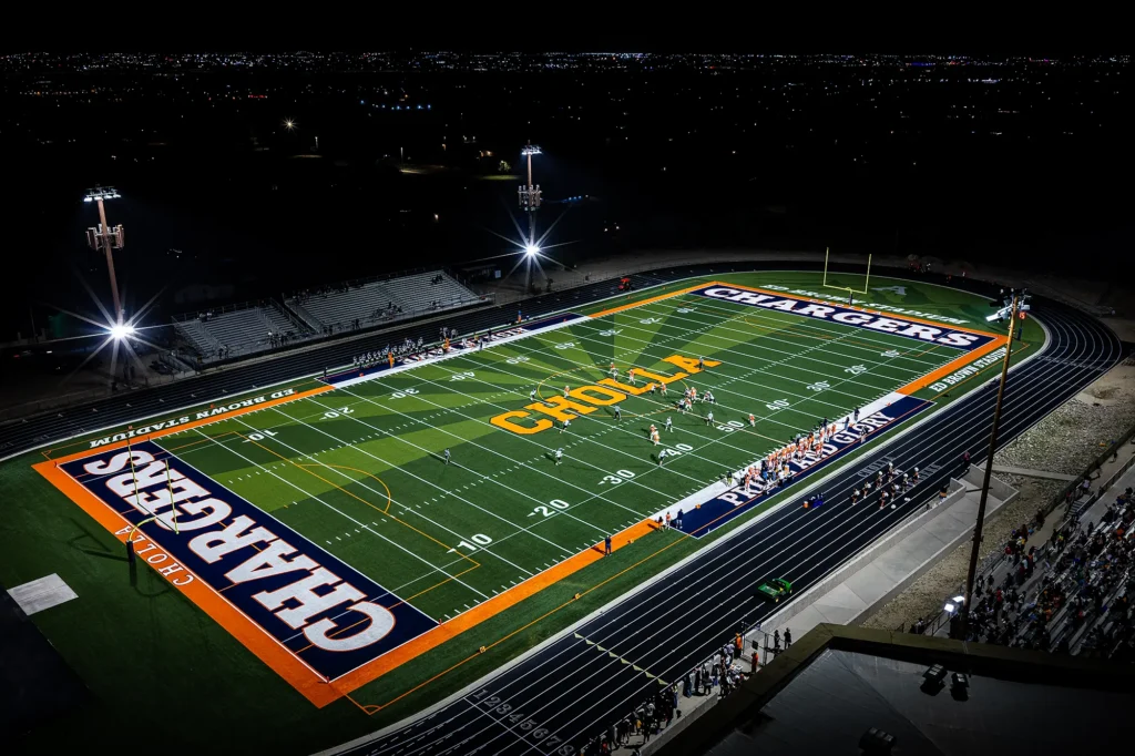 Night aerial view of a football game under the lights on the new MMTH turf field at Cholla High School’s Ed Brown Stadium.