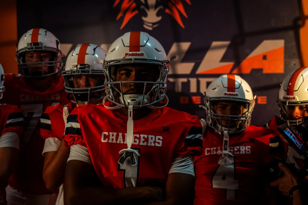 Cholla Chargers football players in orange jerseys and white helmets lined up in front of the Cholla Athletics banner before a game.