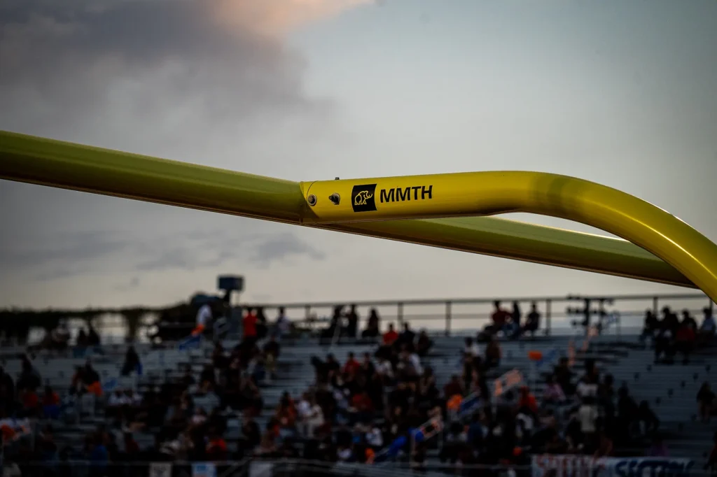 Close-up of a yellow MMTH-branded goal post at Cholla High School, with fans in the stadium bleachers behind it at dusk.
