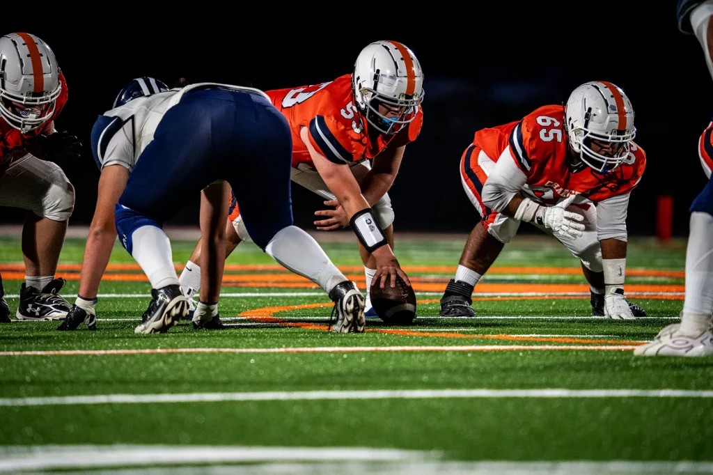 Cholla Chargers linemen in orange uniforms set at the line of scrimmage against an opponent in navy, on the new turf field.