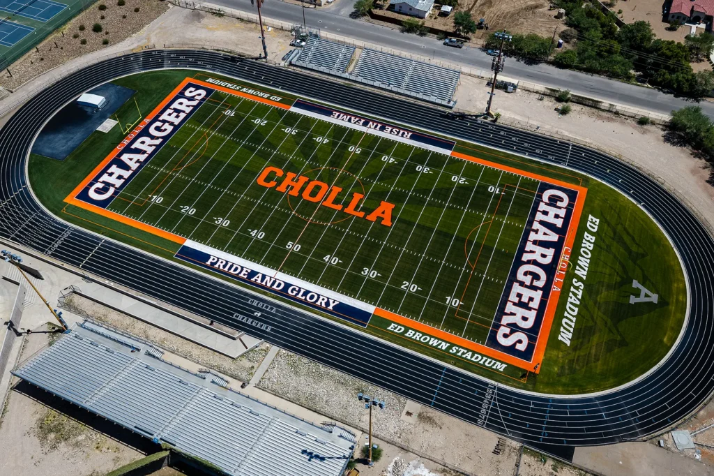 Daytime aerial view of Ed Brown Stadium at Cholla High School, showing the MMTH turf football field with navy and orange end zones, the running track and the bleachers.