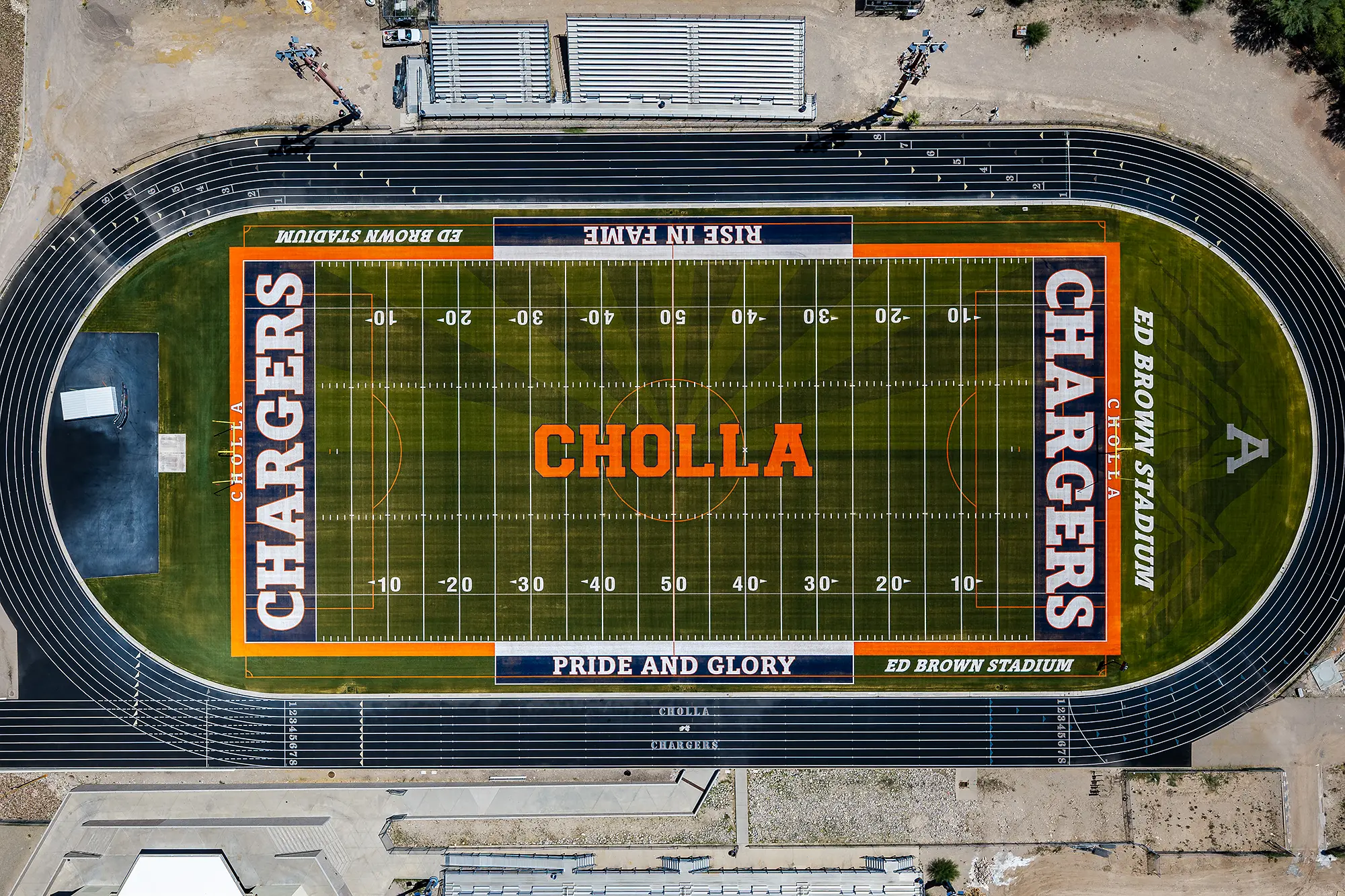 Overhead drone view of the Cholla High School football field, showing “CHARGERS” end zones, the orange “CHOLLA” midfield logo, “PRIDE AND GLORY” and “RISE IN FAME” sideline graphics, and the track around it.