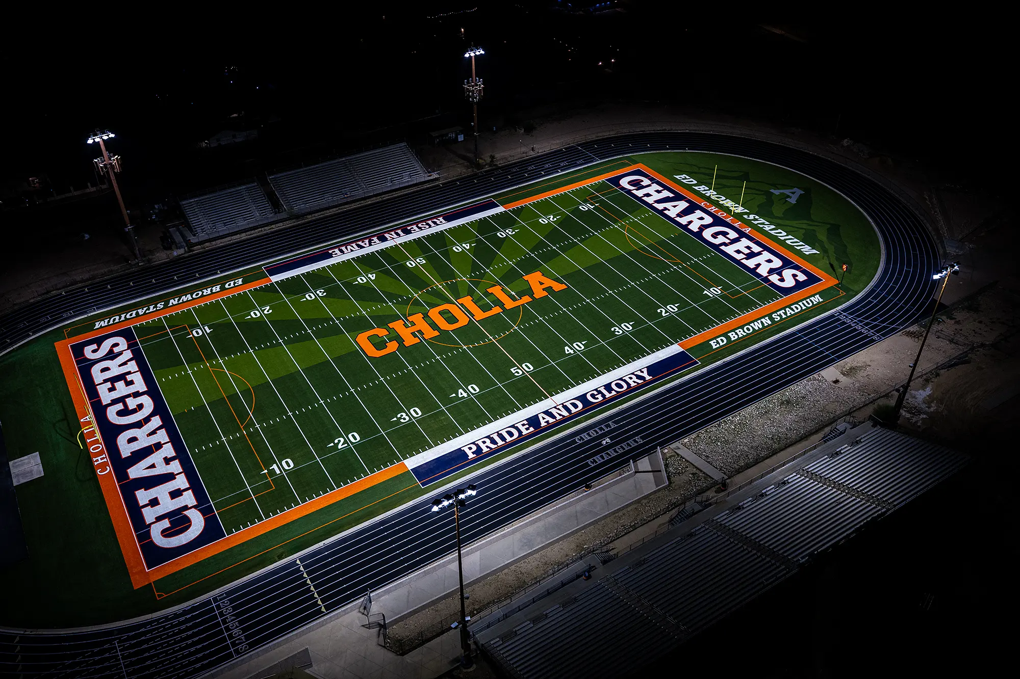 Aerial night view of the lit Cholla High School football field at Ed Brown Stadium, showing the green turf with orange “CHOLLA” at midfield and navy “CHARGERS” end zones, by MMTH