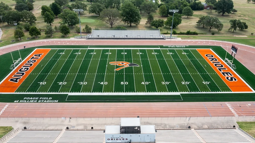 Aerial view of the Augusta High School football field at Poage Field at Hillier Stadium, with orange AUGUSTA and ORIOLES end zones, a midfield Oriole logo, green synthetic turf, a surrounding track, and grandstands