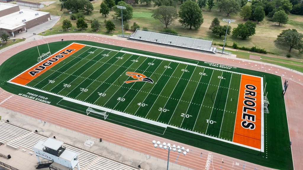 Angled aerial view of Poage Field at Hillier Stadium at Augusta High School, showing the full green turf football field with orange end zones lettered AUGUSTA and ORIOLES, the track, and home bleachers