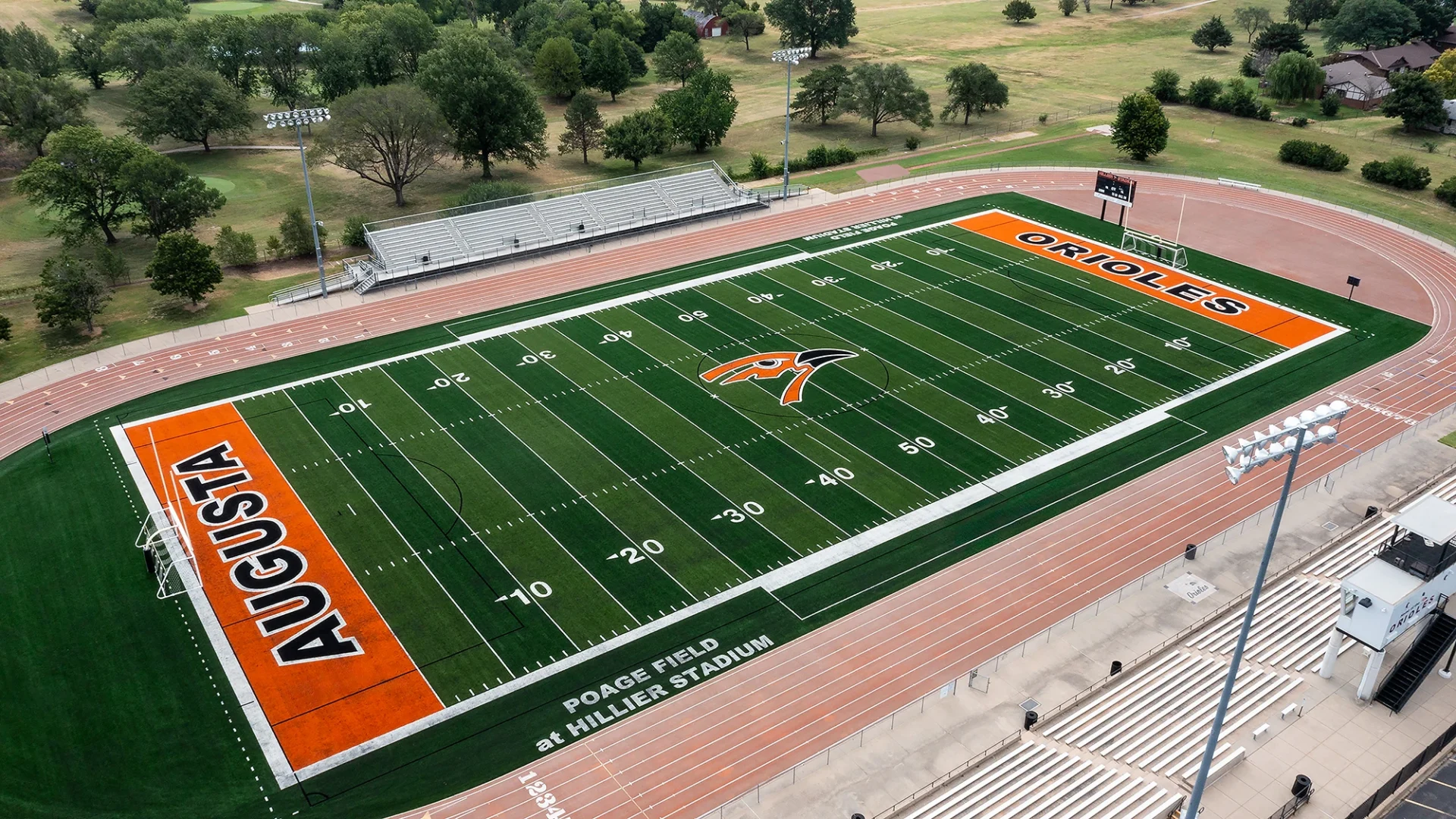 Elevated aerial view of the Augusta High School turf football field at Poage Field at Hillier Stadium, with orange AUGUSTA and ORIOLES end zones, the Oriole logo at midfield, and trees surrounding the stadium