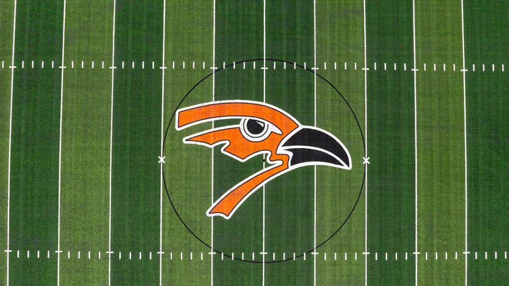 Close-up aerial view of the orange and black Augusta Orioles bird logo at midfield on the green synthetic turf at Poage Field at Hillier Stadium