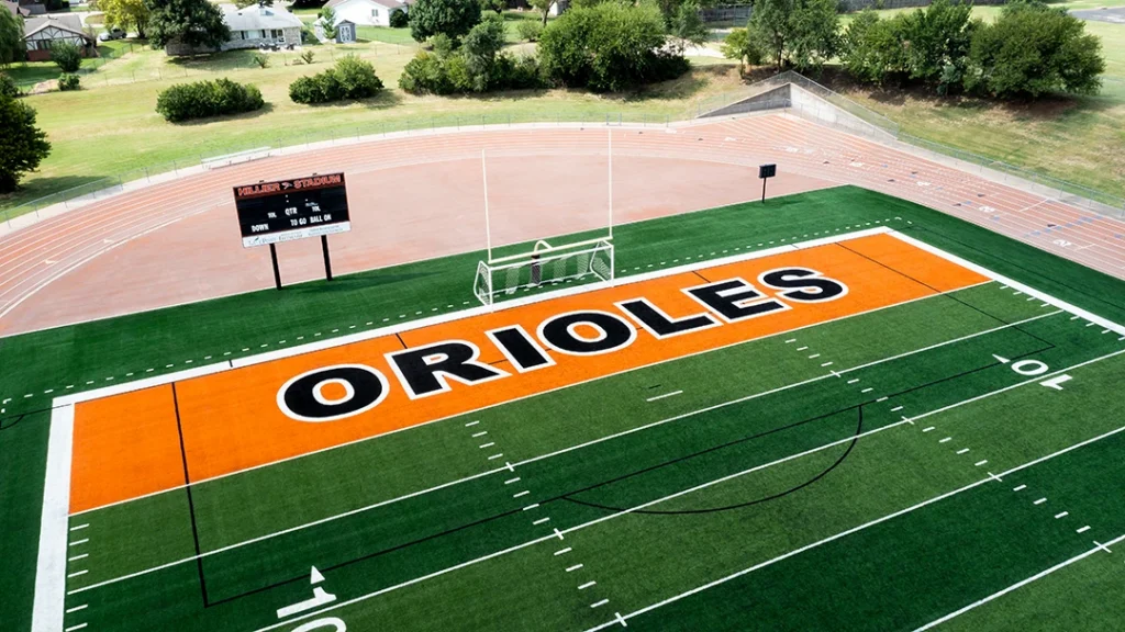 Low aerial view of the orange ORIOLES end zone at Augusta High School's Poage Field at Hillier Stadium, with the goalpost, Hillier Stadium scoreboard, and track in the background